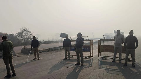 Police officers from UP standing in front of barricades on a road in the fog. one is holding a laathi.