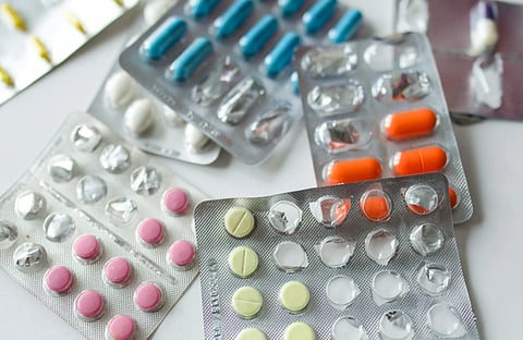 Blister packs of various pills and capsules are scattered on a white surface. The pills are pink, yellow, orange, and blue, indicating variety.