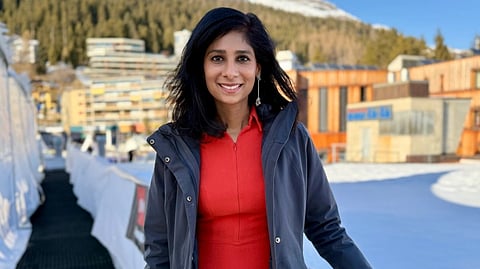 Harvard Professor  Gita Gopinath in a red dress and gray coat stands outdoors, smiling. Snow covers the ground, with modern buildings and a forested mountain in the background under a clear blue sky.