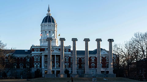 Front view of a historic building with a central dome, flanked by six tall stone columns. A person with a dog stands in the grassy foreground.