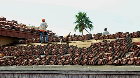 Workers on a roof with stacked terracotta tiles, under a clear sky. A palm tree is visible in the background.