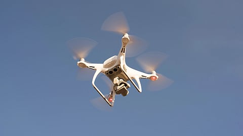 A white drone with four propellers hovers mid-air against a clear blue sky. The propellers are in motion, creating a dynamic and airy feel.