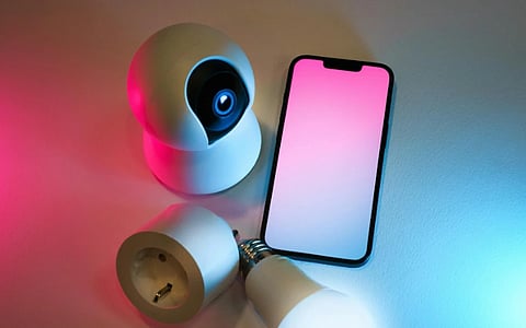 A smartphone with a pink gradient screen is surrounded by a white security camera, smart plug, and LED bulb, all under blue and red lighting. Modern tech vibe.