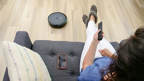 A woman relaxes on a gray sofa, holding a TV remote. A robot vacuum cleans the wooden floor. Her phone rests beside her, conveying a cozy, modern atmosphere.