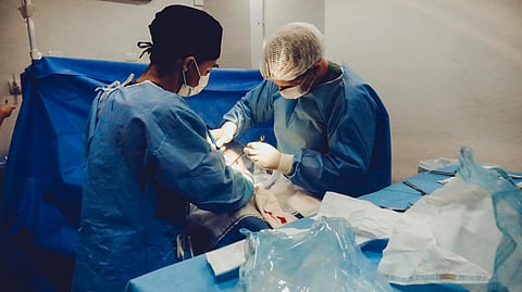 Two surgeons in blue scrubs and masks perform surgery under bright lights in a sterile operating room.