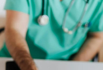 A person in green medical scrubs, with a stethoscope around their neck, reaches for a tube labeled "MASC." A jar of pills and a clipboard are on the desk.
