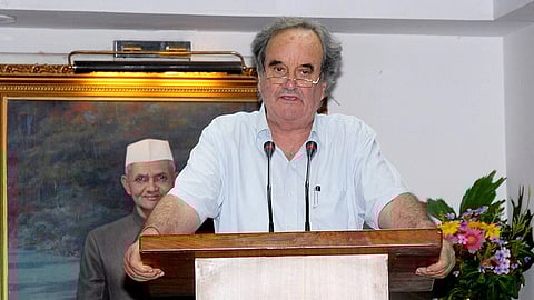 Sir Mark Tully speaking at a podium, in an interior hall, at an Indian Railways seminar, a portrait of former Indian PM Lal Bahadur Shashtri ji behind him