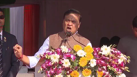 Himanta Biswa Sarma mid-speech, speaking into a mic at a podium covered in flowers while raisng his hand.