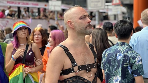 A person wearing a leather outfit stands confidently in a colorful Pride parade. Behind them, participants in rainbow attire and vibrant hats celebrate.