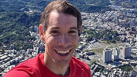 American climber Alex Honnold in a red shirt takes a selfie from the top of Taipei 101 with the background showing a sprawling cityscape with lush hills and a bright, cloudy sky.