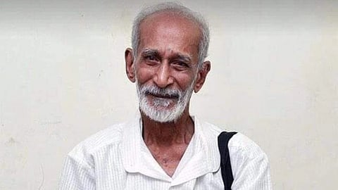 Kalyanasundaram, with white hair and beard, smiling gently, wearing a white shirt and dhoti, holding a black bag, standing against a plain wall.