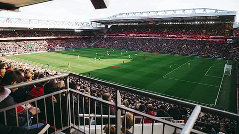 A packed stadium with fans watching a soccer match. Players in red and white uniforms compete on a sunlit green field. 