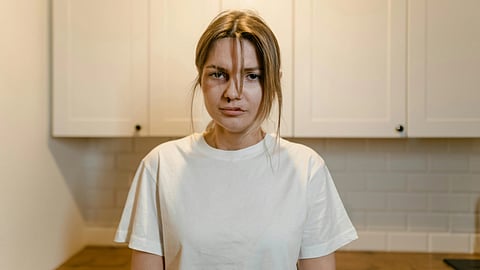 A woman in a white t-shirt stands in a kitchen with a neutral expression. Light-colored cabinets and a tiled backsplash form the background.