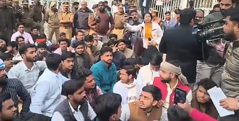 A large group of people sits on the ground, surrounded by police and journalists with cameras. The mood appears tense and focused.
