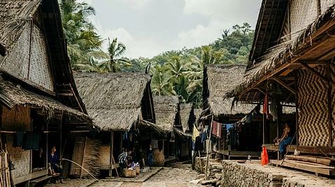 A narrow village path flanked by traditional thatched-roof huts, with clothes hanging to dry. Palm trees and hills are visible in the background, creating a serene and rustic atmosphere.
