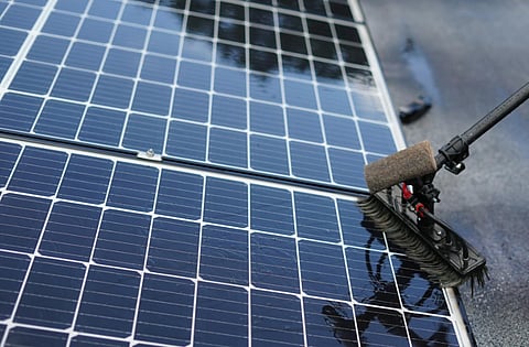 A worker uses a brush with a long handle to clean shiny solar panels, reflecting light. The scene conveys maintenance and sustainability efforts.