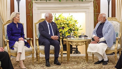 PM Modi European Commission Ursula von der Leyen and European Council President António Costa seated in ornate chairs in a formal setting, with vibrant yellow and blue floral arrangements. They are engaged in conversation, creating a warm, diplomatic atmosphere.
