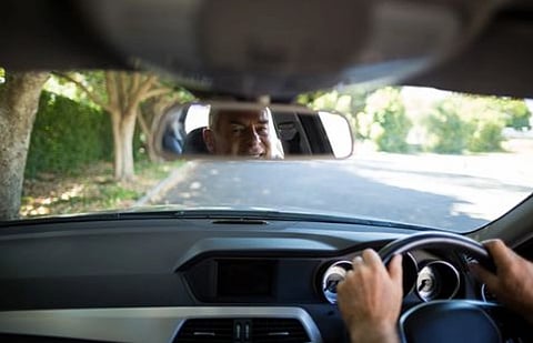 View from inside a car, focusing on the dashboard and steering wheel. A person's reflection is visible in the rearview mirror, driving on a sunny road lined with trees.