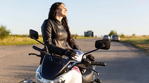 A woman in a leather jacket stands next to a bike on the road while smiling.