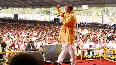 Himanta Sarma on stage in a yellow kurta, speaking into a mic with his hadn raised in front of a crowd.
