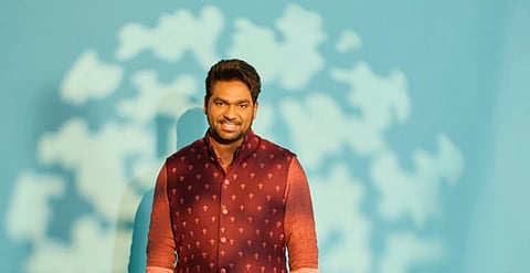 A smiling man in a maroon patterned vest stands against a blue background. Light creates abstract leaf shadows, adding a playful, relaxed feel.