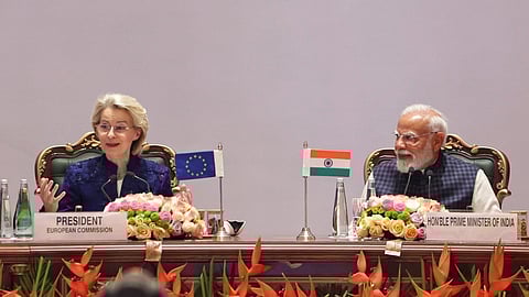 PM Modi and Ursula von der Leyen sits at a table during a diplomatic meeting. The European Commission President gestures, while the Indian Prime Minister listens, both smiling.