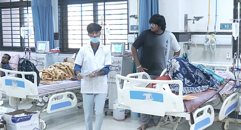 A hospital room with three patients resting in beds, covered with blankets. A nurse in a mask is walking by, and a visitor stands nearby. The atmosphere is calm.