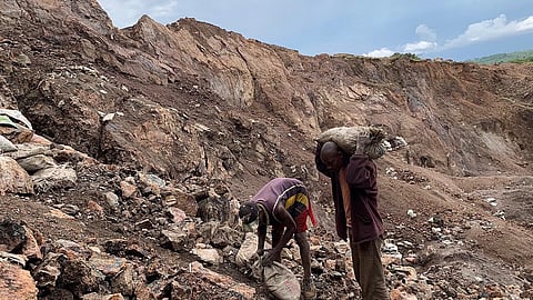 Two individuals work in a rocky quarry, one carrying a heavy sack on his shoulder, the other bent over collecting stones. The scene is overcast and industrious.