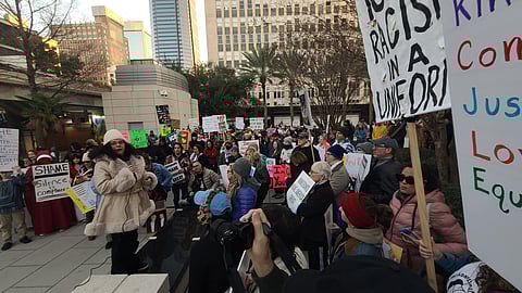 A large crowd gathers at a protest in a city square, holding diverse signs promoting equality and justice. The atmosphere is energetic and unified.