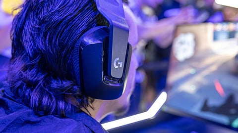 Person with long hair wearing large headphones and intensely gaming at a computer in a vibrant, crowded setting, glowing with blue neon light.