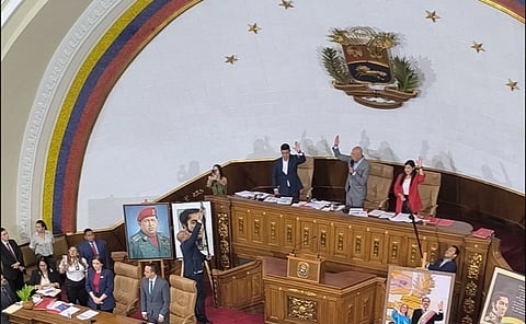 A group of people stand in a large assembly hall with a high, curved ceiling and a colorful emblem. Portraits of notable figures are displayed. The atmosphere is formal.