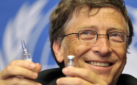 Close up shot of Bill gates holding syringe and medicine in his hands. in the image he is seen smiling and looking towards his right side. 