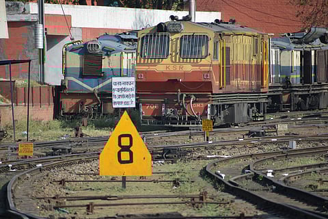 A red-and-yellow KSR train engine is parked on railway tracks alongside a blue engine. A prominent yellow triangular sign with the number eight is in the foreground, with additional signage displaying text in a local language. The setting appears to be a station or railway yard on a sunny day.