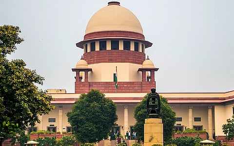 Image of Central Wing of the Supreme Court of India where the Chief Justice's courtroom is situated. A large building with a prominent dome, featuring the Indian flag. In the foreground, there's a statue surrounded by greenery.