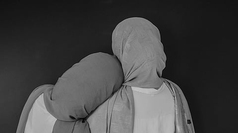 Two people wearing headscarves stand back-to-back, one resting their head on the other’s shoulder. Both wear light clothes, conveying warmth and support.