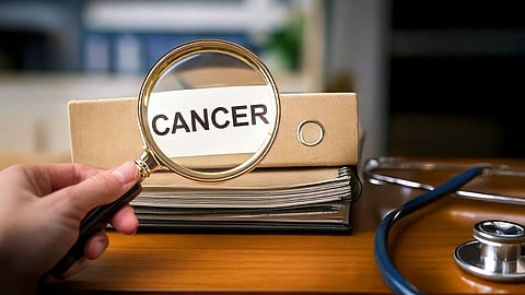 A magnifying glass held over a file labeled "Cancer" on a desk. A stethoscope lies beside it, suggesting medical research or diagnosis focus.