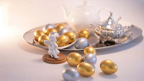 Silver and gold eggs on a tray with a porcelain teapot and ornate sugar bowl. A small white angel figurine sits in front, creating an elegant scene.