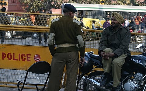 A police officer in uniform stands near another policeman sitting on a motorcycle behind a yellow barricade labeled "POLICE" on a busy street. 