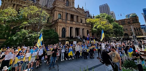 Large crowd gathered outside a historic building, holding Ukrainian flags and protest signs. The mood is determined, with individuals of diverse backgrounds.