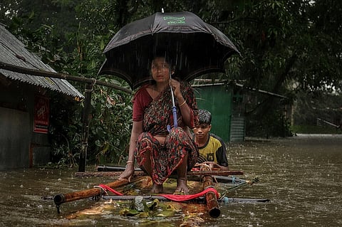 A woman crouches on top of a makeshift raft with an umbrella being pushed by a man in a flooded street.