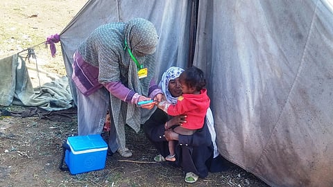 Two women stand with a small child in front of a tent, while one injects the child with a vaccine.