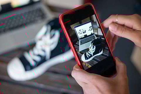 Hands holding a smartphone with a red case, taking a photo of a black and white sneaker. A laptop displays the sneaker image in the background.