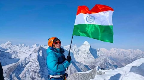 Arunachal Pradesh mountaineer Kabak Yano in a blue jacket and orange helmet triumphantly holds the Indian flag atop a snowy peak, with vast, snow-covered mountains under a clear blue sky.