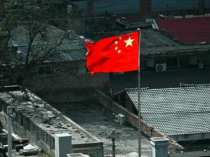 A vibrant red Chinese flag with yellow stars waves on a rooftop against an urban backdrop of gray buildings and trees under a clear sky.