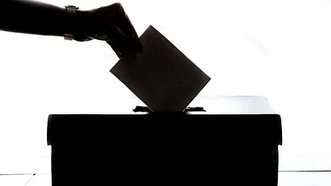 A silhouetted hand places a ballot into a voting box against a bright white background, conveying themes of democracy and civic responsibility.