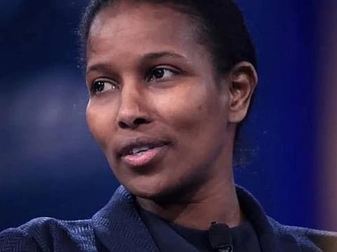 Image of  Writer Ayaan Hirsi Ali speaking on stage, wearing a dark, textured blazer. The background is blurred blue and purple, 