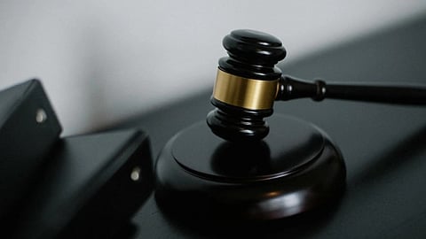 A wooden judge's gavel with a brass band rests on a dark surface, symbolizing law and justice. Nearby, blurred black binders suggest legal documents.