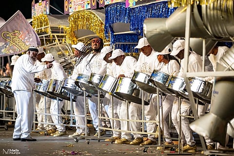 A lively steel band dressed in white performs energetically on stage, surrounded by colorful decorations and shimmering gold and blue tinsel, creating a festive atmosphere.