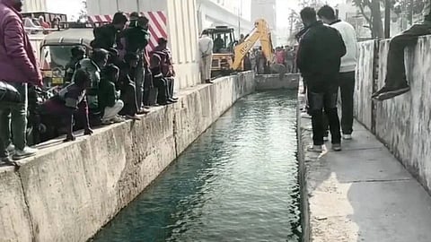 People gather along a concrete canal with clear water, watching a backhoe loader working nearby. The scene conveys curiosity and anticipation.
