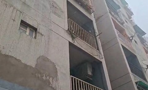 A multi-story building viewed from below, featuring weathered concrete walls and balconies with metal railings. The sky is overcast, adding a somber tone.